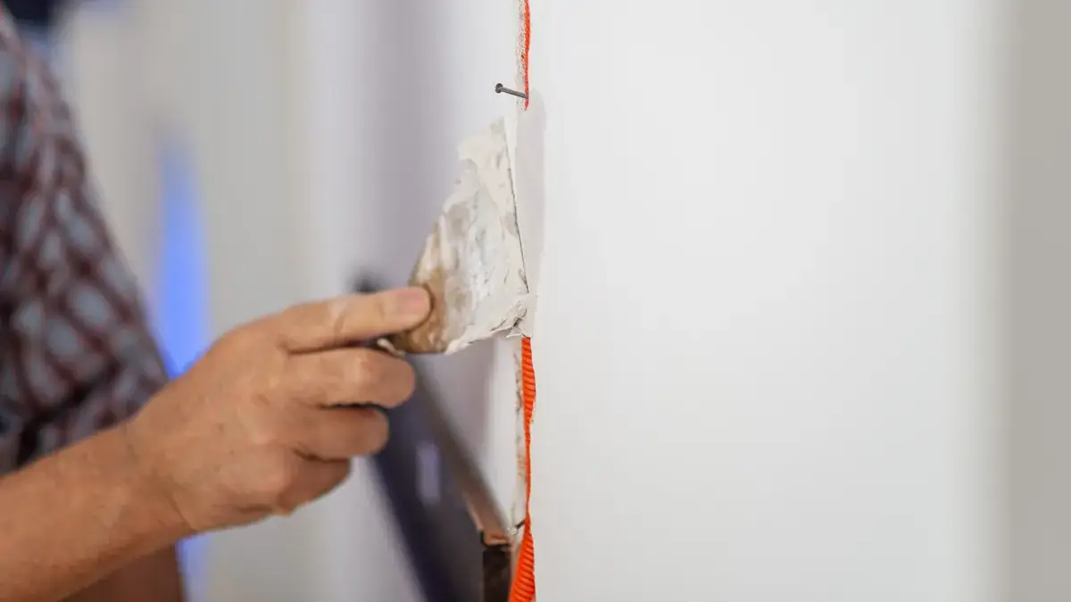Hand applying jointing compound over orange fibreglass mesh jointing tape and a nail head on a plasterboard wall