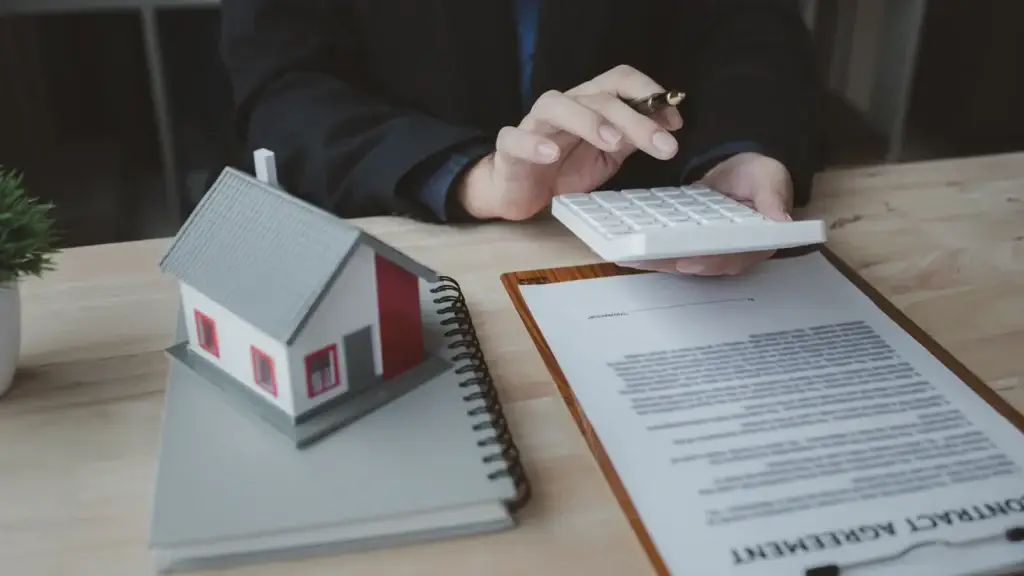 Person using a calculator next to a contract agreement document and house model on a wooden desk
