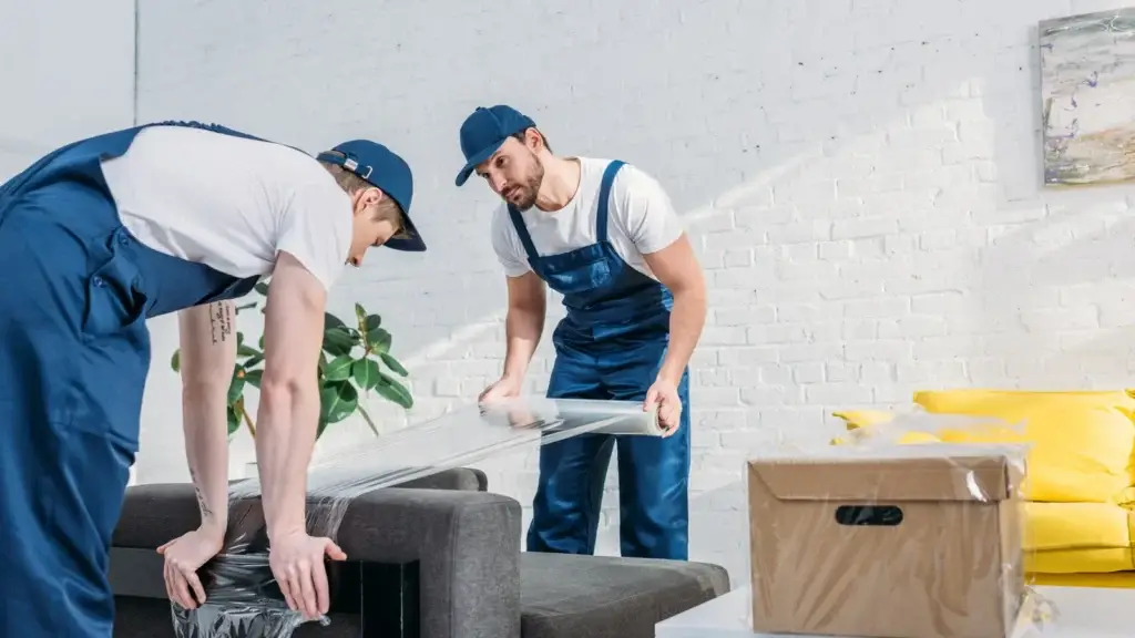 Two removalists in blue uniforms wrapping a dark couch in protective plastic wrap in a residential living room before a house move