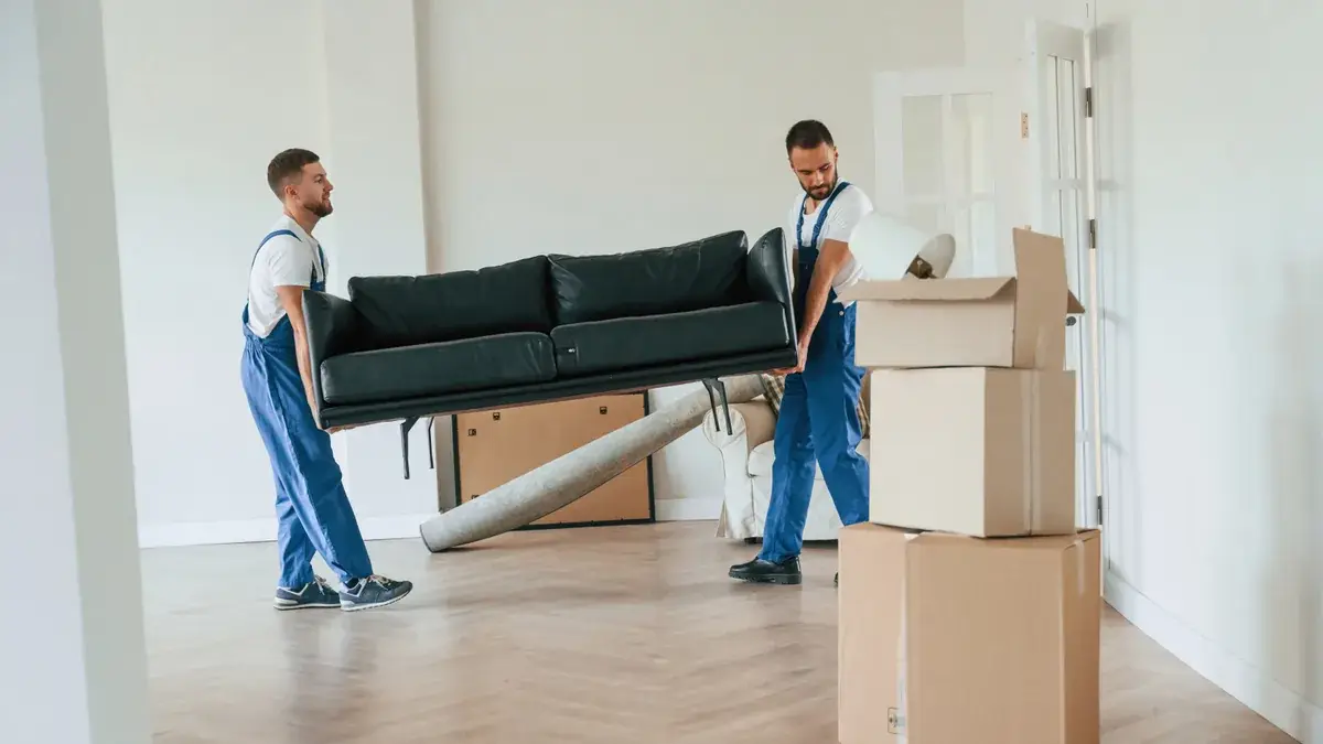 Two removalists in blue dungarees carrying a black leather couch through an empty room with herringbone floors and moving boxes during a house move