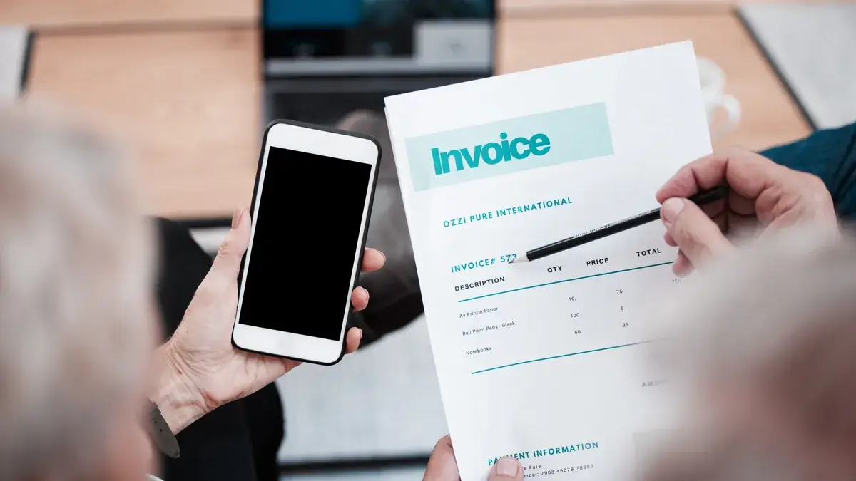 Person holding a phone and reviewing a printed invoice with a pen, checking the supplier name and details