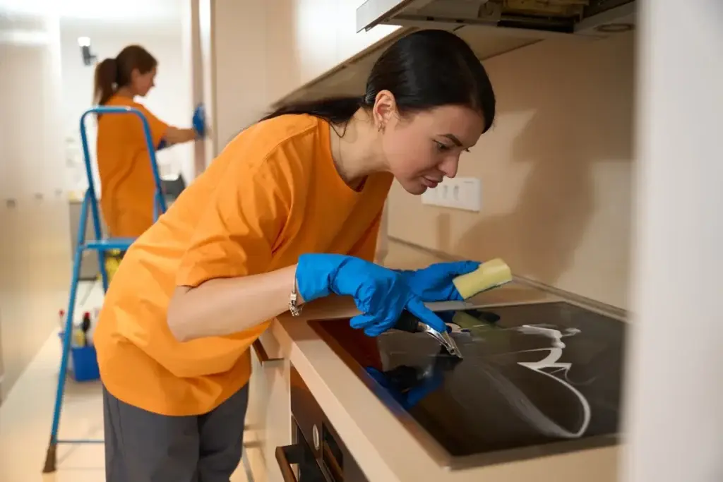 Professional cleaner in orange uniform scrubbing a glass cooktop with sponge