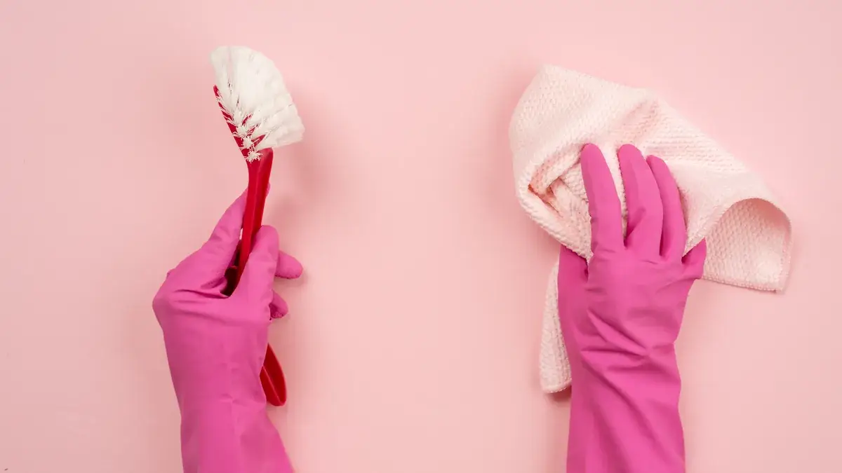 Two gloved hands in pink rubber gloves holding a scrubbing brush and a cleaning cloth against a pink background
