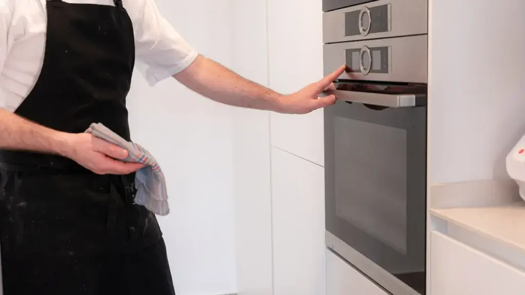 A cleaning professional in an apron inspecting a built-in oven in a modern white kitchen during an end of lease clean