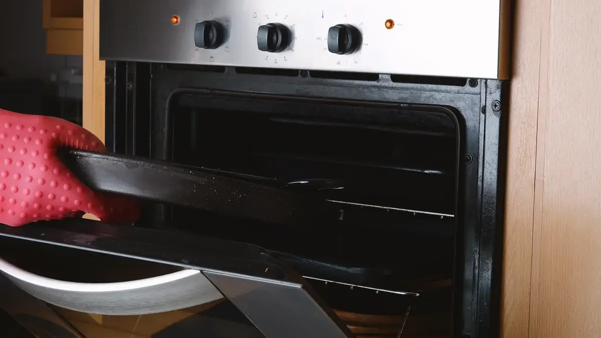 A gloved hand removing a soiled baking tray from a heavily used oven with visible grease buildup and baked-on residue