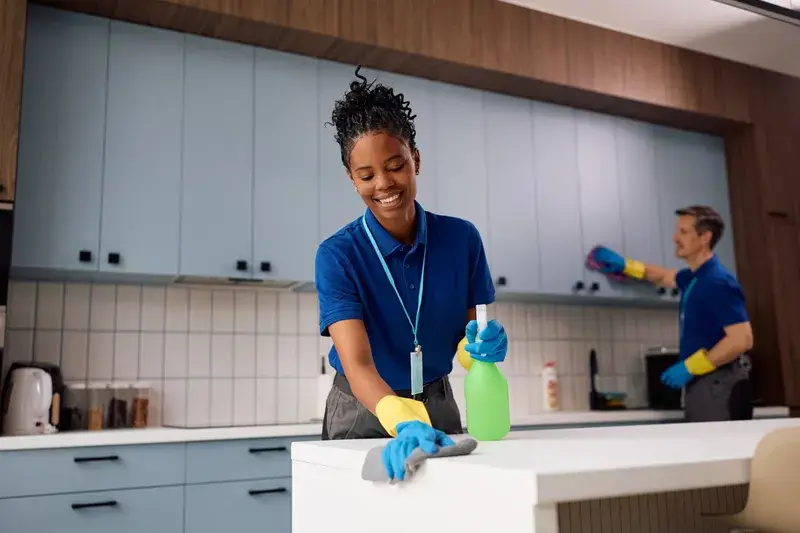 Two verified independent end of lease cleaners working together in a modern Australian kitchen, with one cleaner spraying and wiping a white countertop while a second cleaner wipes upper cabinet shelves in the background, with professional equipment visible in a realistic local service setting