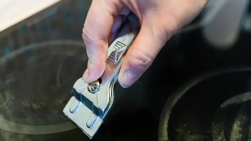 Hand using a chrome cooktop scraper at a low angle on a black glass-ceramic cooktop surface with cooking zone rings