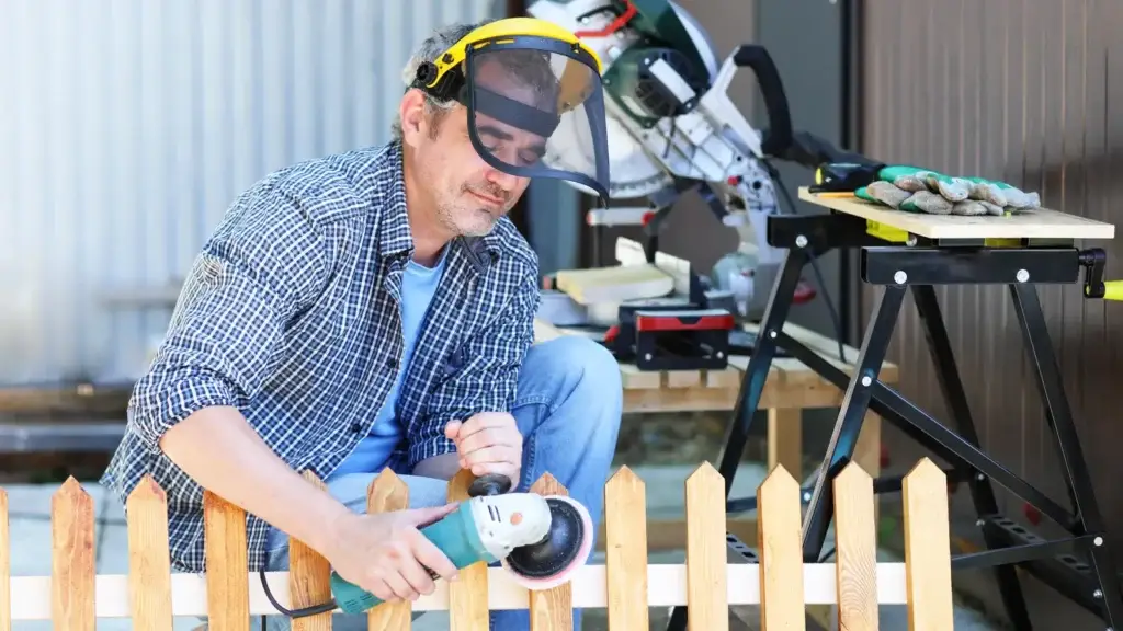 Handyman using an angle grinder on a timber picket fence outdoors, wearing a yellow face shield