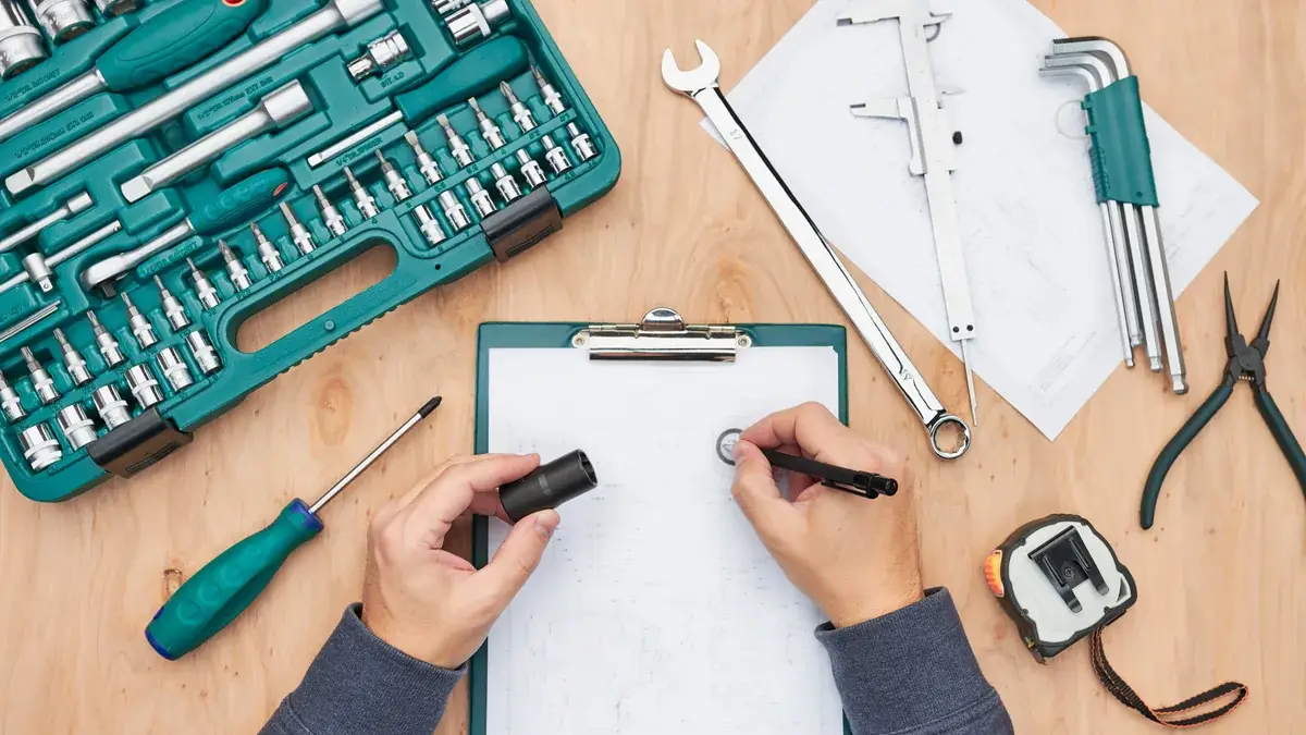Overhead view of hands writing on a clipboard surrounded by a socket set, wrenches and hand tools on a timber surface