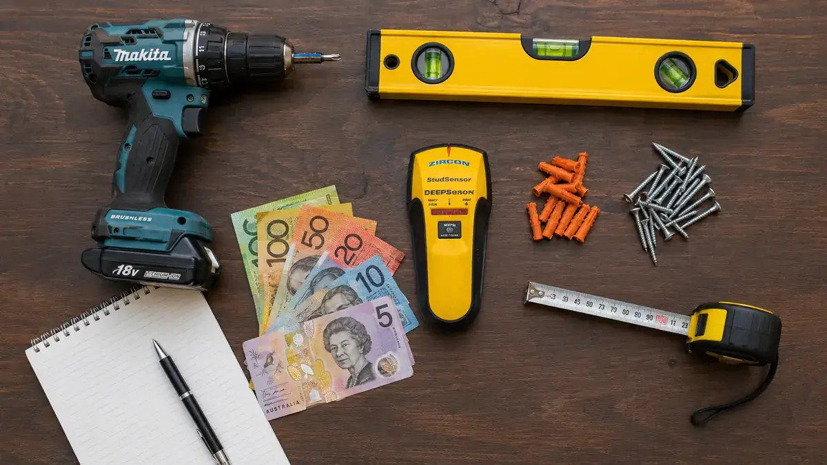 Makita drill, spirit level, stud finder, tape measure, wall plugs, screws, notepad and Australian banknotes on a wooden surface