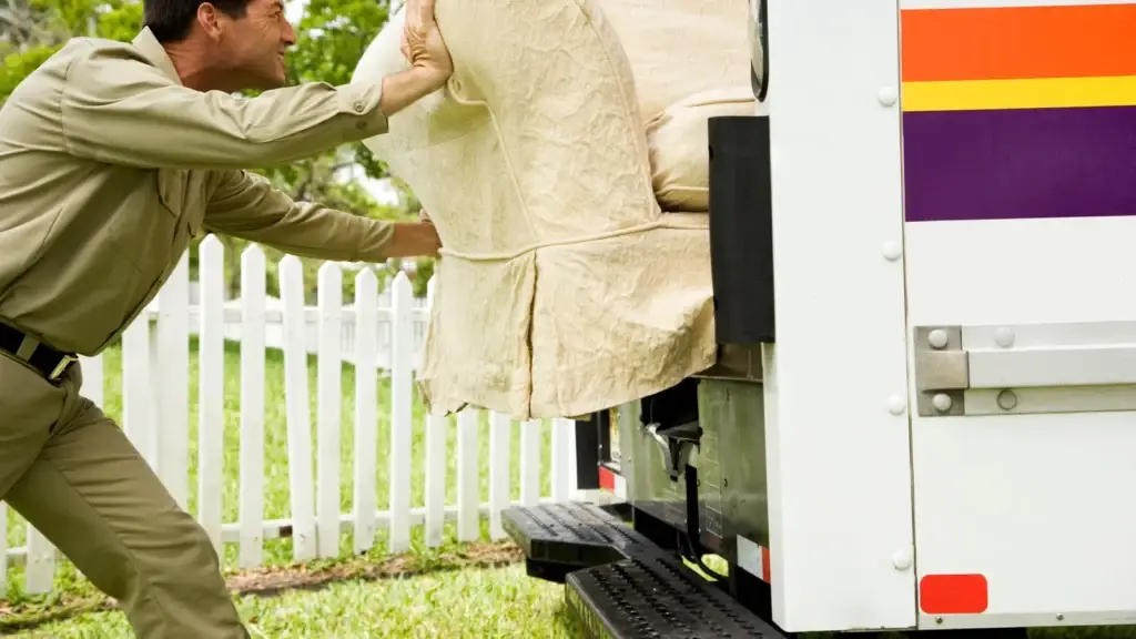 Removalist in uniform loading a blanket-wrapped armchair onto a removal truck ramp outside a residential property