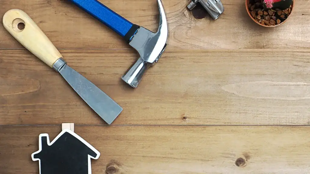 Home improvement tools including a hammer and scraper beside a house icon on a timber surface