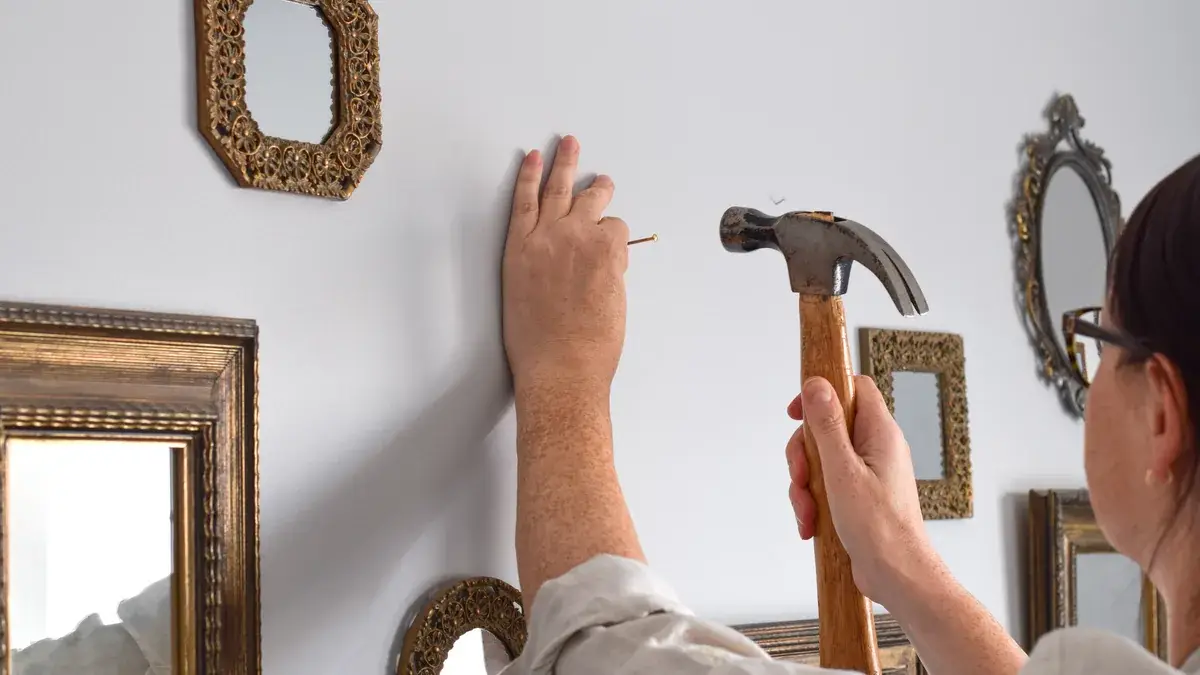 Woman using a hammer to nail a picture hook into a white wall surrounded by multiple decorative mirrors