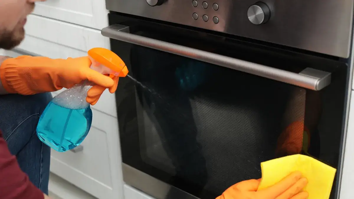 Man in orange rubber gloves spraying cleaning product onto the oven door glass while wiping it with a yellow cloth during an end of lease clean