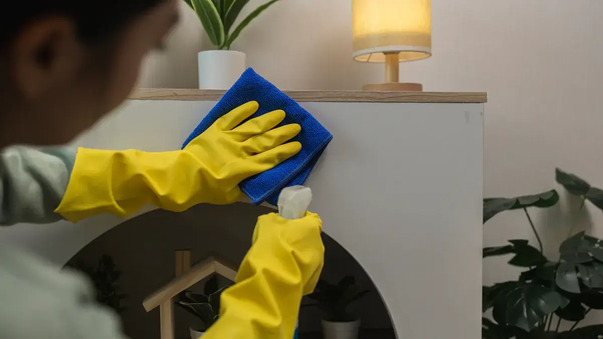 Person wearing yellow rubber gloves wiping a white furniture surface with a blue microfibre cloth during a house clean