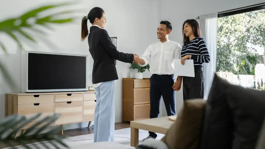 A real estate agent shaking hands with a smiling couple in a furnished Australian rental property living room.
