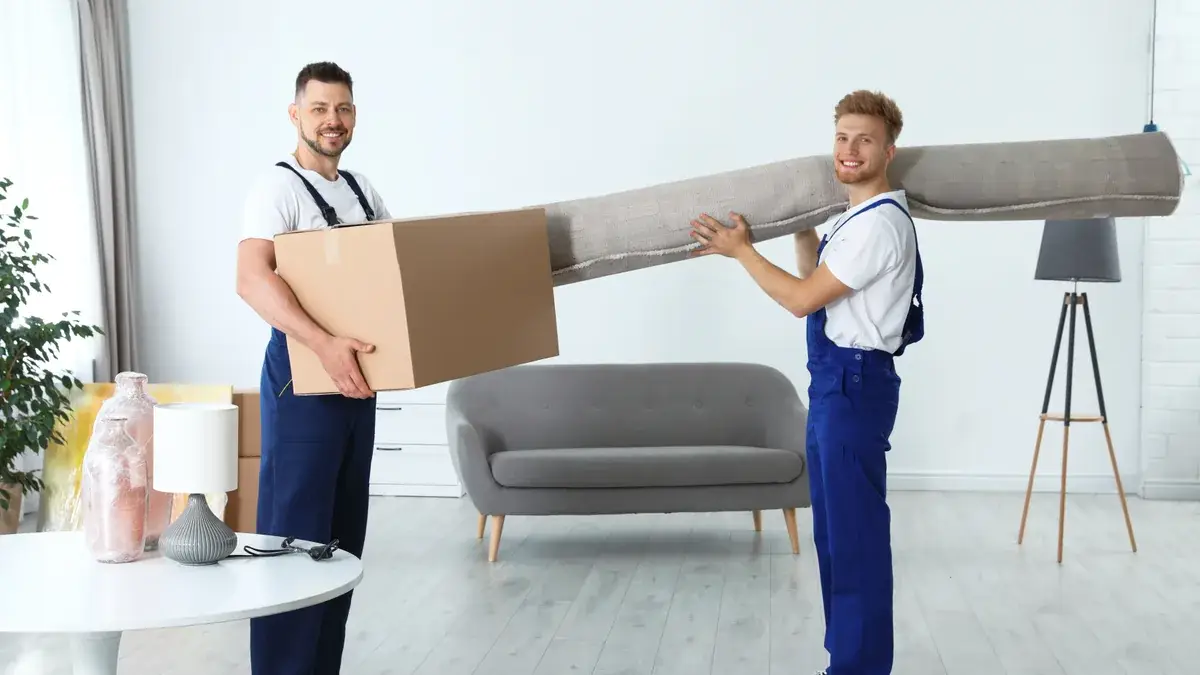Two removalists in blue overalls carrying a cardboard box and a rolled carpet inside a lounge room