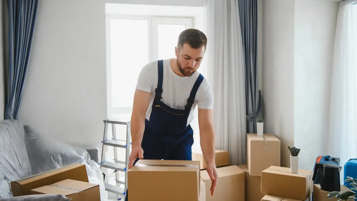Professional removalist in blue overalls lifting a cardboard box in a modern lounge room surrounded by packed moving boxes
