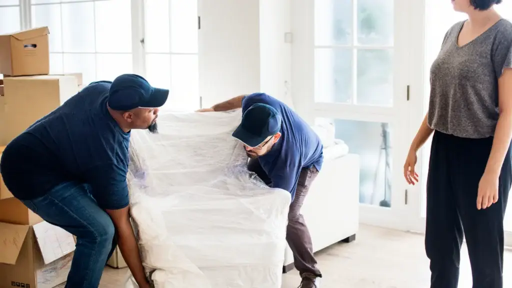 Two removalists lifting a plastic-wrapped armchair past stacked moving boxes while a woman watches