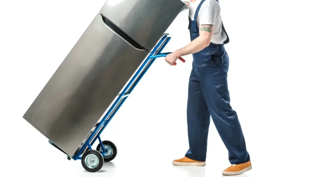 Removalist in blue work overalls using a blue appliance trolley to move a stainless steel top-freezer fridge