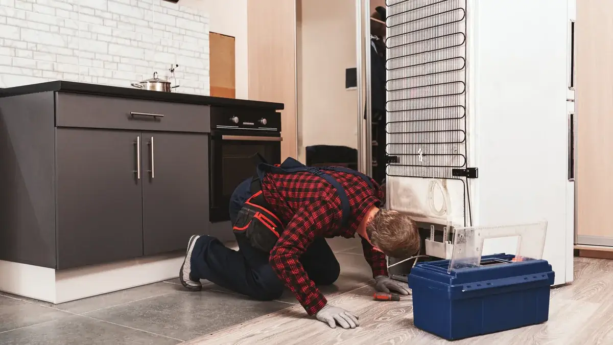 Technician kneeling on a kitchen floor inspecting the condenser coils at the back of a fridge pulled away from the wall