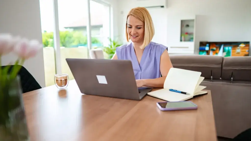 Woman smiling while working on a laptop at a dining table in a bright open-plan home, with a notebook and phone beside her