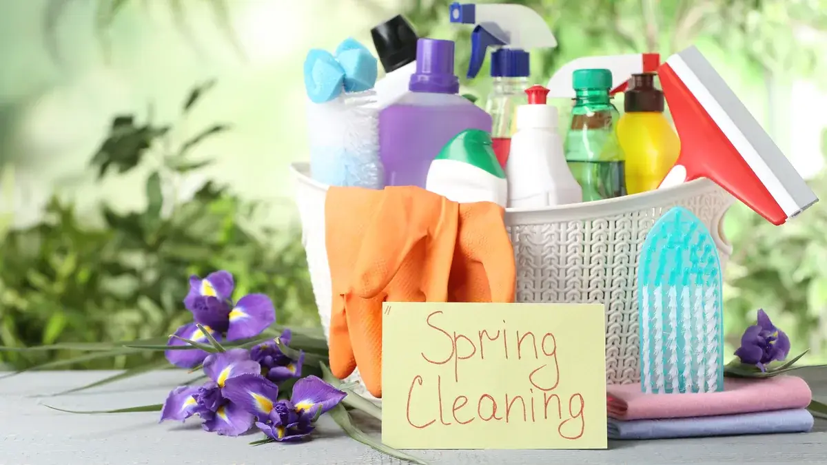 Spring cleaning supplies including spray bottles and rubber gloves in a basket with a Spring Cleaning sign and flowers