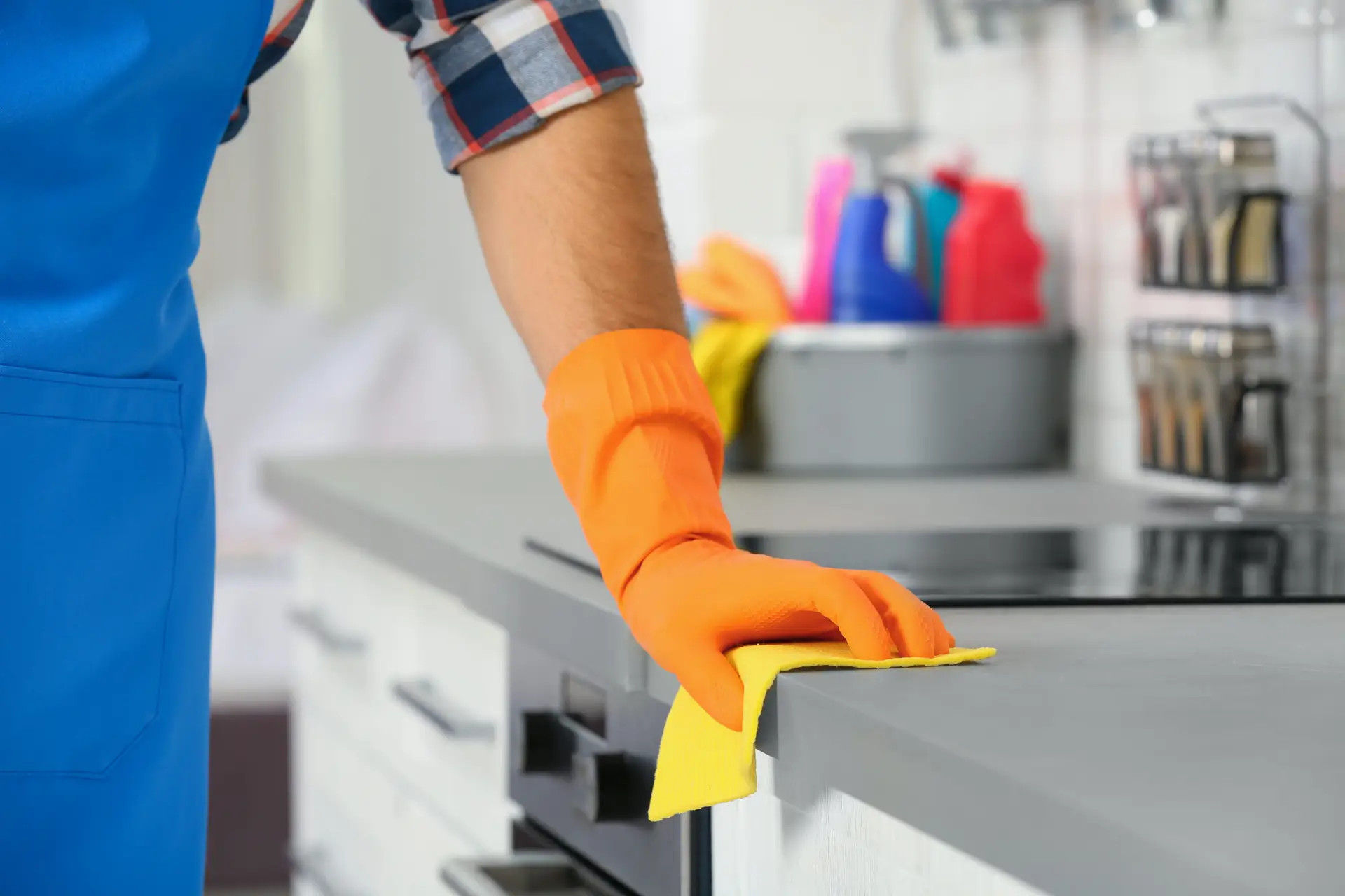 An Urgent Pro cleaner wiping down every surface of a Brisbane rental kitchen — benchtops, cupboard doors, the splashback tiles and the front of the oven — in warm Queensland afternoon light with a cleaning caddy sitting on the kitchen floor