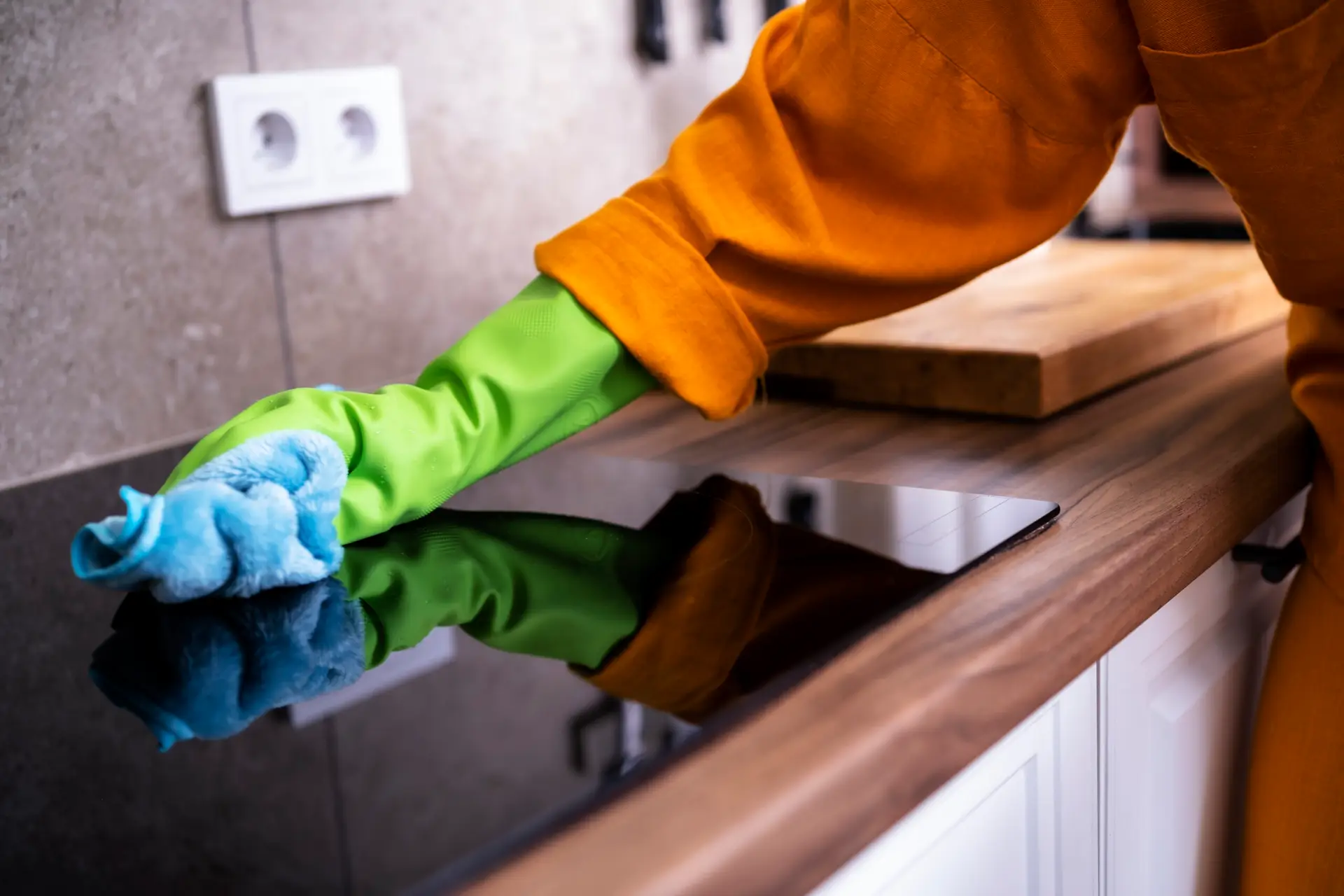 An Urgent Pro cleaner wiping down the stone benchtop, the sink and the splashback of a modern Gold Coast rental kitchen in bright Queensland coastal light, with a cleaning caddy on the floor and the oven door open beside them ready for the next cleaning task