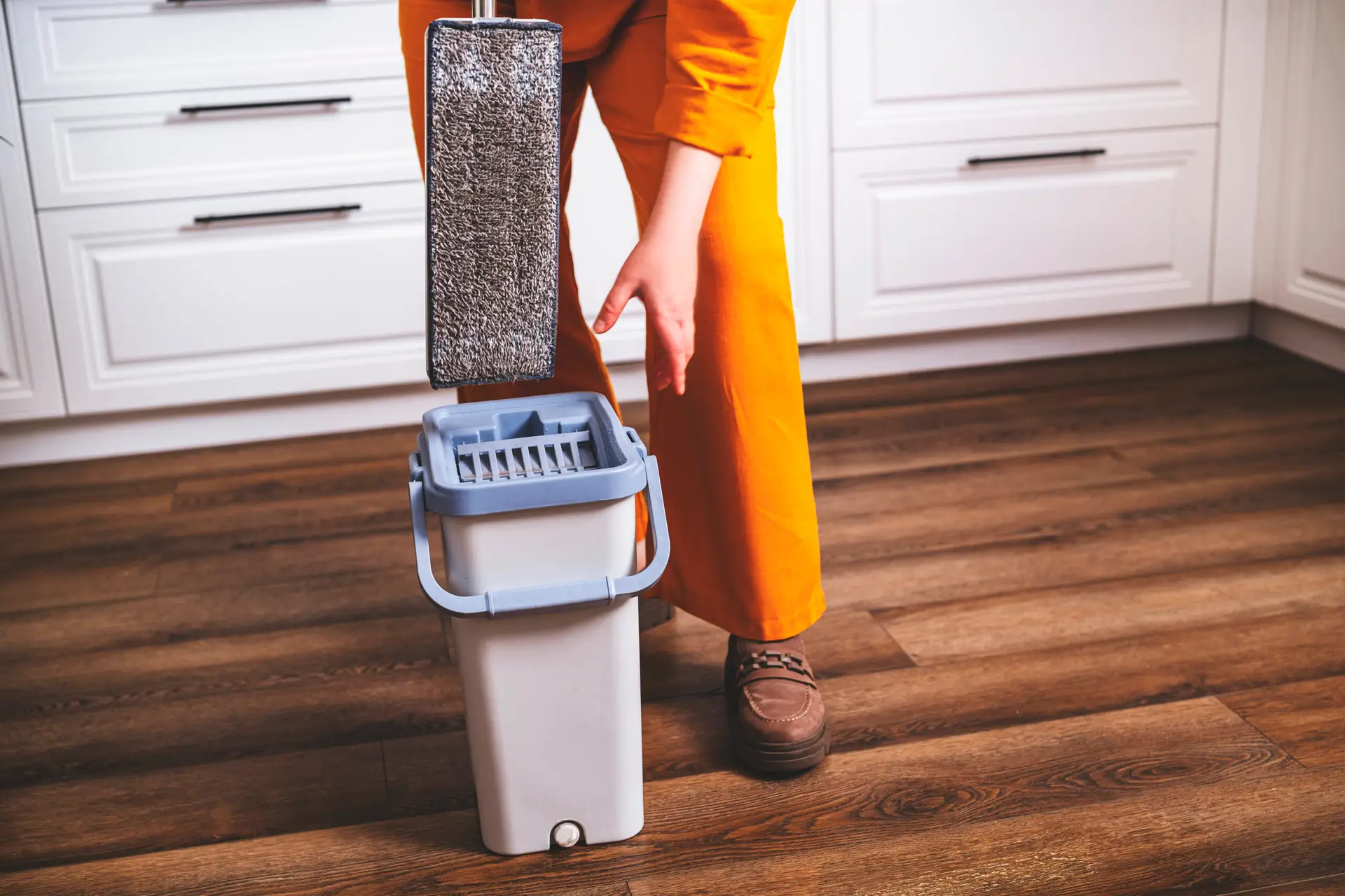 An Urgent Pro cleaner sweeping and mopping the large light-coloured tiled floor of an empty Gold Coast rental living room, working from the back of the room toward the sliding door that opens to a balcony with coastal Gold Coast visible in the background