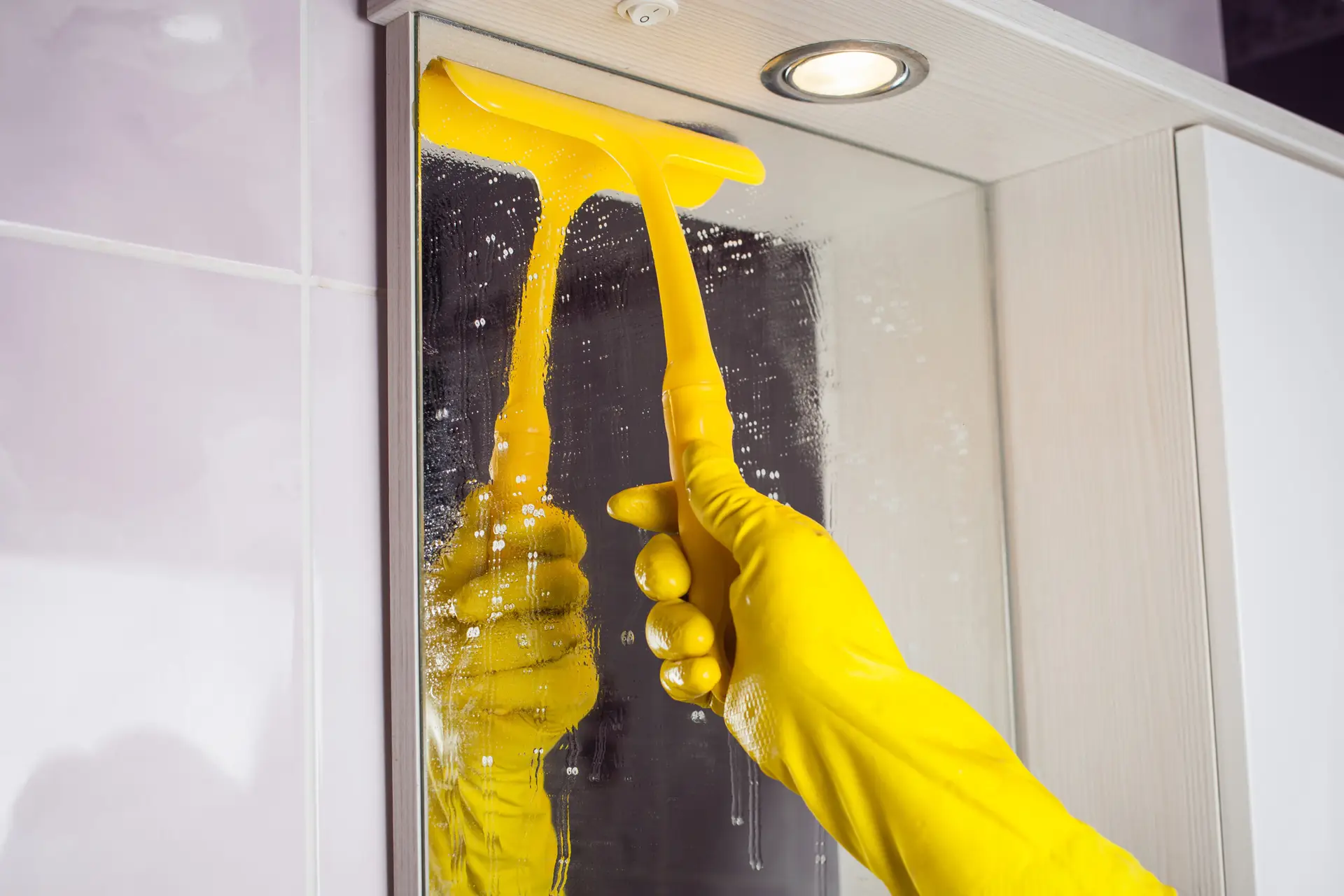 An Urgent Pro cleaner in a Melbourne rental bathroom polishing the vanity mirror with a glass cleaner, while the basin, tapware and the tiles around it are freshly cleaned and drying — with the cleaning products lined up along the vanity edge