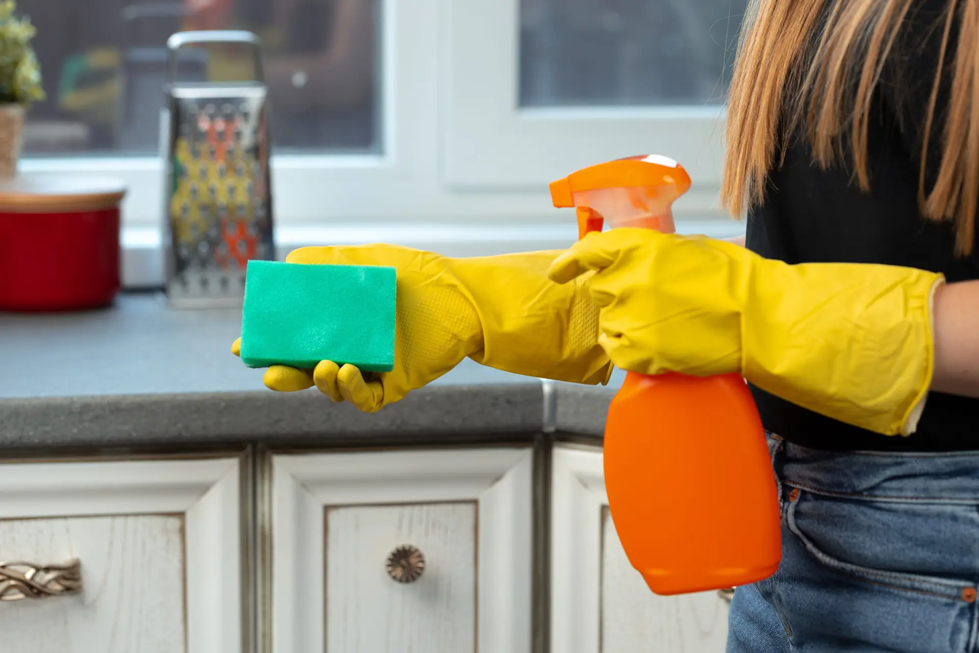 An Urgent Pro cleaner in a Melbourne rental kitchen cleaning the full length of the benchtop including the sink, stovetop surround and cupboard handles, using a spray cleaner and a yellow microfibre cloth in a modern rental interior