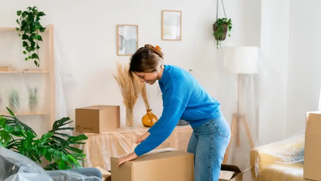 Woman sealing a cardboard moving box in a lounge room with shrink-wrapped furniture in the background